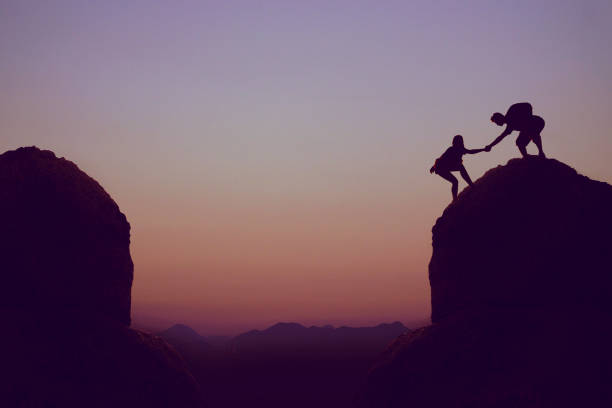 Silhouette of a person helping another up a steep rock side against the backdrop of a purple sunset